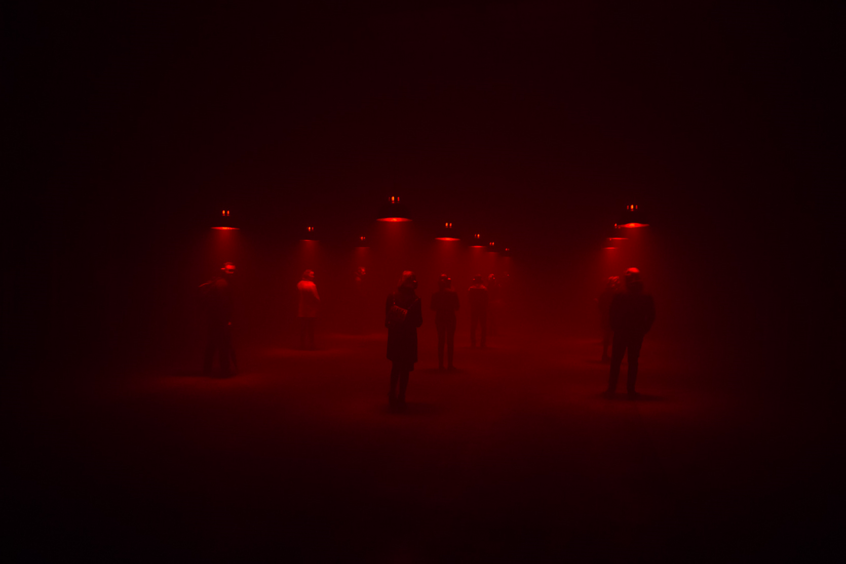 People stand under individual red lights, listening in a dimly lit room