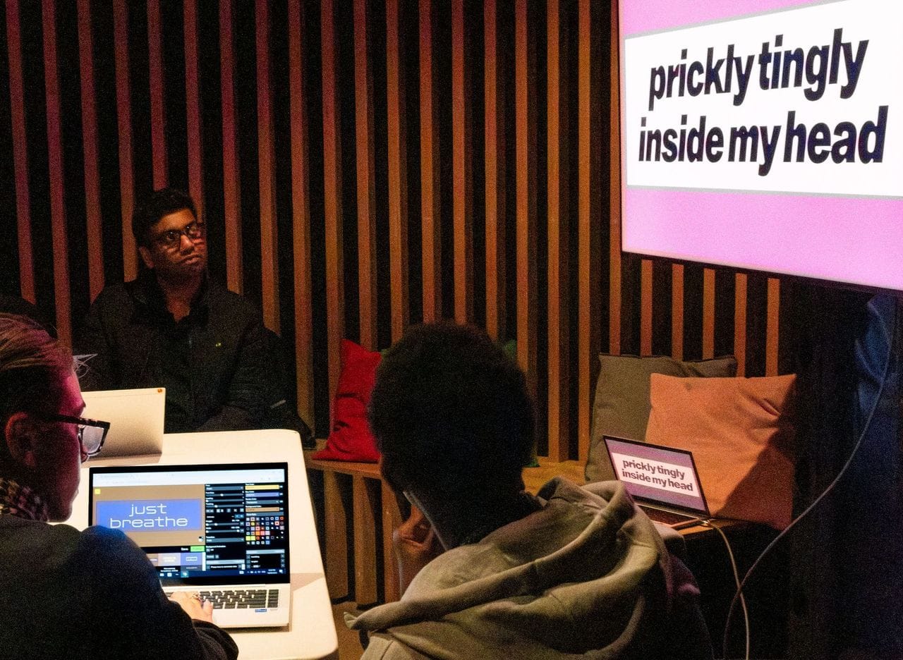 In a warmly, dimly lit room walled by vertical strips and angled benches of timber, a bespectacled brown-skinned man looks pensively up at a large TV screen. On that screen, a white box floats in a bubblegum pink background, and bold black text reads 'prickly tingly inside my head’. As the eye follows a dangling cable falling from behind the screen, it lands on a half-folded laptop on which the same words and colours can be seen. Closer to the viewer, the backs of two other people’s heads are also turned toward the big TV, but one of these people’s fingertips rest on the keyboard of a laptop with an app open, showing a similar layout – coffee coloured background, pale blue text box and white text reading: just breathe.