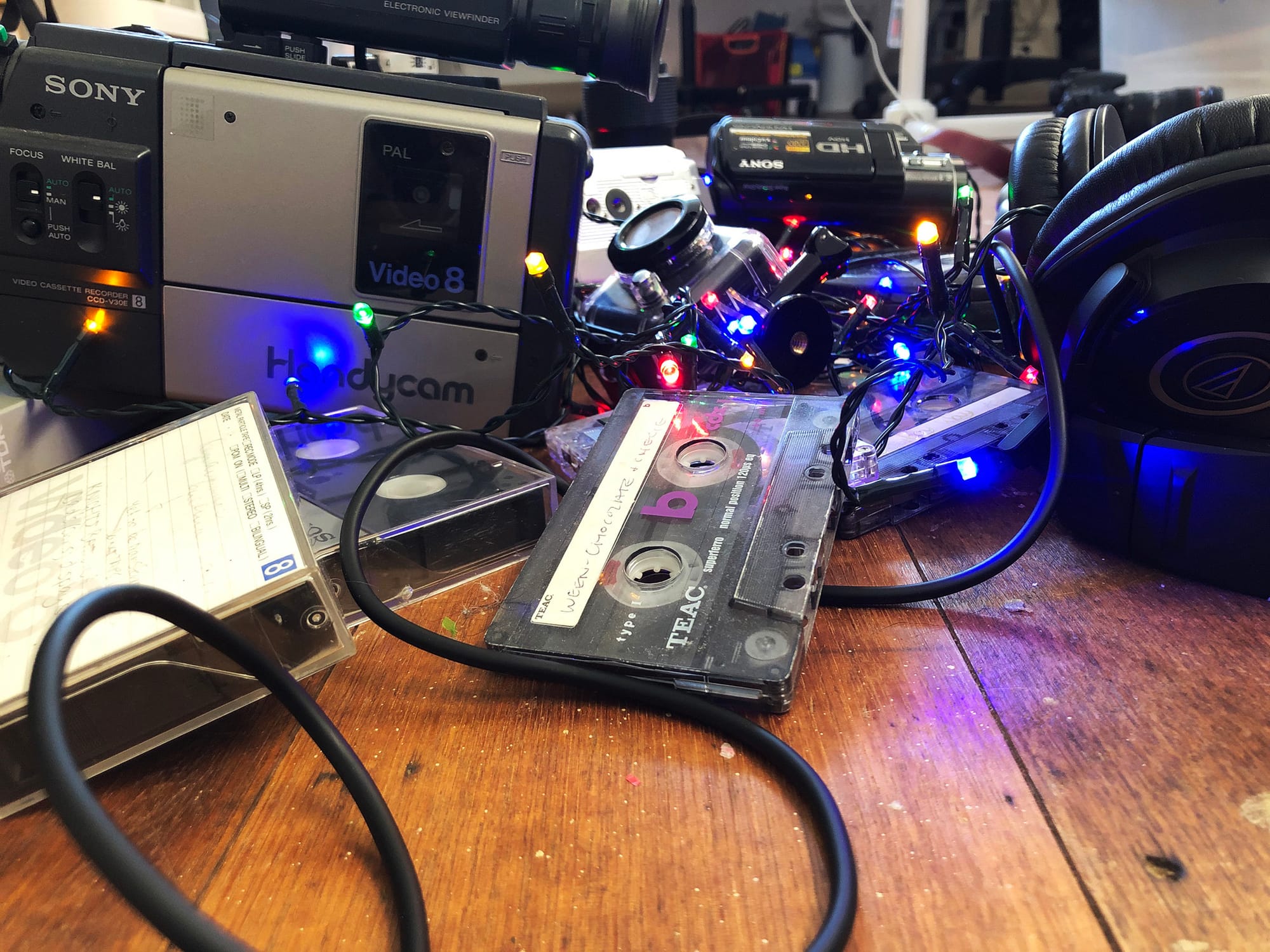 A jumble of old cassettes, VHS tapes, and video equipment on a wooden table with LED party lights on top